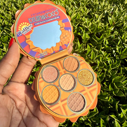 The palette features six waffle-patterned pans in warm shades of orange, brown, and gold glitter, with a mirror on the inside of the orange lid.